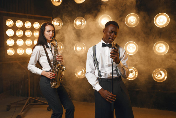 Mężczyzna jazzman i kobieta saksofonista z saksofonem na scenie z reflektorami. Jazzowi wykonawcy grający na scenie - Zdjęcie, obraz