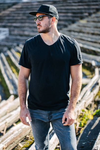 Hipster handsome male model with beard wearing black blank t-shirt and a baseball cap with space for your logo or design in casual urban style - 写真・画像