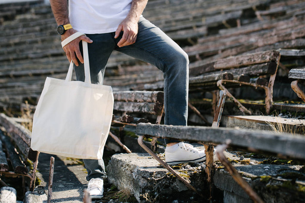 Young man holding white textile eco bag against urban city background. Ecology or environment protection concept. White eco bag for mock up. - 写真・画像
