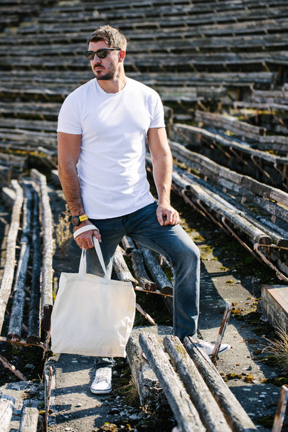 Молодий чоловік тримає білий текстильний еко-пакет на фоні міста. Екологія або концепція захисту навколишнього середовища. Біла еко сумка для сміху вгору
. - Фото, зображення