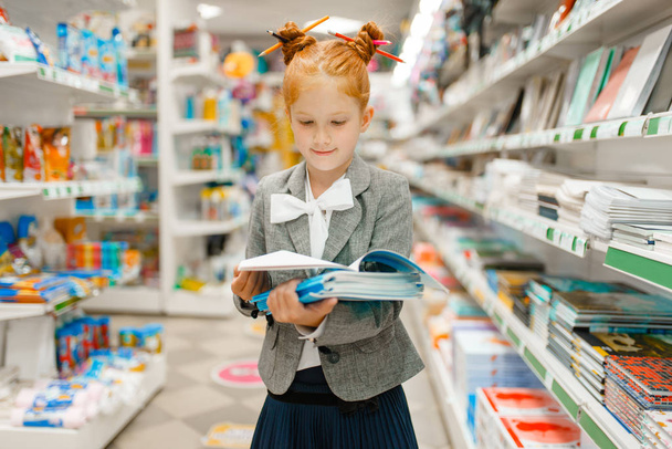 Маленька школярка з блокнотами в канцелярському магазині. Жінка-дитина купує канцелярське приладдя в магазині, школярка в супермаркеті
 - Фото, зображення