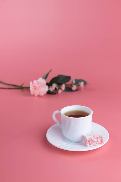 A white cup of tea with a piece of lokum, turkish delight on the side, with delicate flowers on a pink background. Afternoon tea concept - Photo, Image