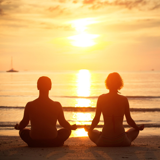 Prática de ioga: um jovem casal sentado na praia do mar na posição de lótus ao pôr do sol
. - Foto, Imagem