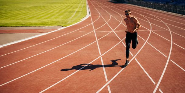 Horisontaalinen kuva aktiivisesta lenkkeilijästä juoksemassa radalla stadionilla. Urheilullinen mies hölkkäämässä aurinkoisena päivänä stadionilla. Ihmiset, terveelliset elämäntavat ja urheilun käsite
 - Valokuva, kuva