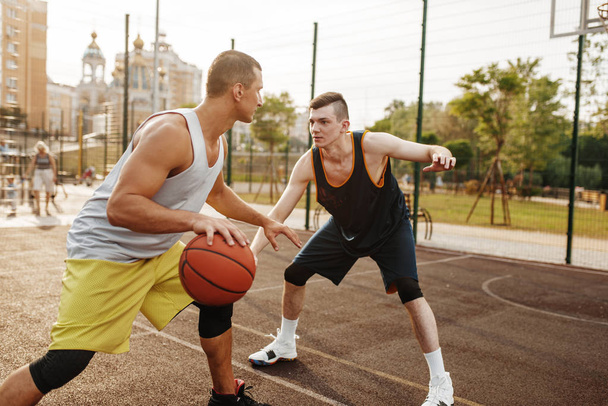 Two basketball players playing intense match on outdoor court. Male athletes in sportswear play the game on streetball training - Zdjęcie, obraz