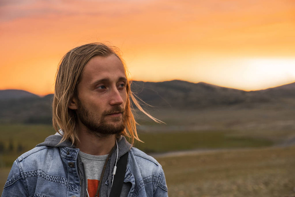 A man looks away at sunset in the mountains. The traveler is wearing a denim jacket. Blond with blue eyes and a dark beard. Backpacker in a hike in the summer mountains. Mountain Altai. Portrait. - 写真・画像
