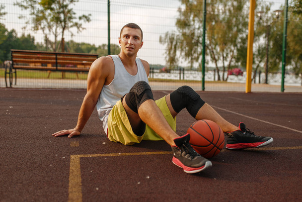 Jugador de baloncesto con pelota sentado en el suelo en la cancha al aire libre. Atleta masculino en ropa deportiva descansando después del entrenamiento de streetball
 - Foto, Imagen