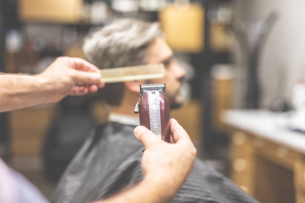 Professional barber holding comb and clipper, client sitting in armchair in the background. - Photo, Image