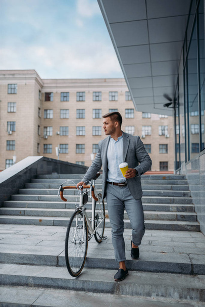 Businessman in suit with bicycle comes down the stairs at the office building in downtown. Business person riding on eco transport on city street, urban style - 写真・画像