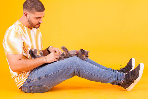 Молодий чоловік потирає котячий живіт в студії на жовтому фоні
 - Фото, зображення