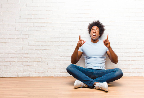young black man looking shocked, amazed and open mouthed, pointing upwards with both hands to copy space sitting on the floor at home - Photo, image