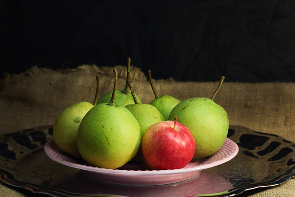 Green organic pears and one small red apple, different not like others, on delicate pink plate on silver metal tray, picturesque still life on coarse retro cloth canvas. Dark atmospheric fall harvest - Photo, Image
