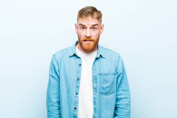 young red head man looking puzzled and confused, biting lip with a nervous gesture, not knowing the answer to the problem against soft blue background - Photo, Image