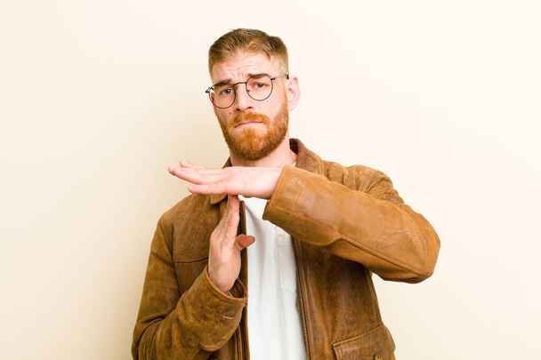 young red head man looking serious, stern, angry and displeased, making time out sign against beige background - Фото, изображение