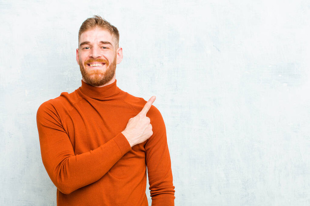 young red head man wearing turtle neck smiling cheerfully, feeling happy and pointing to the side and upwards, showing object in copy space against concrete wall - 写真・画像