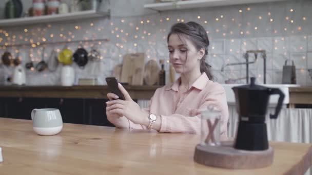 Fiatal nő ül a konyhaasztalánál otthon dolgozik a kis üzletén egy okos telefonnal. Készletfelvétel. Egy fiatal hölgy portréja, amint lehallgatja a mobilját.. - Felvétel, videó