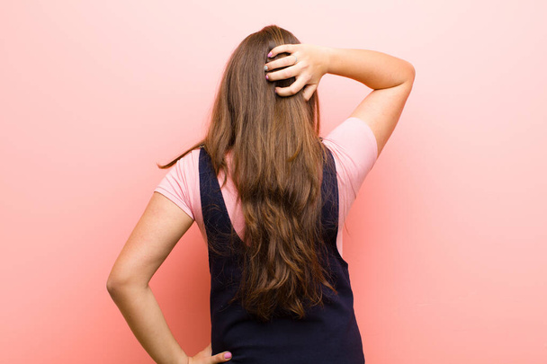 jonge vrouw denken of twijfelen, krabben hoofd, gevoel verward en verward, rug of achteraanzicht tegen roze achtergrond - Foto, afbeelding