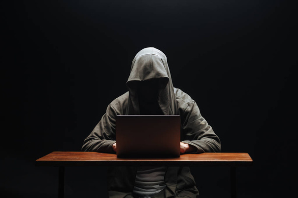 Hacker without a face in a hood with a laptop on a black backgro - Фото, изображение