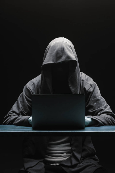 Hacker sem rosto em um capuz com um laptop em um backgro preto
 - Foto, Imagem