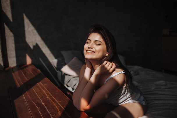 Chica sentada en la cama junto a la ventana a la luz del sol
 - Foto, imagen