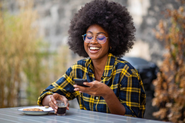 Portrait de jeune femme afro-américaine assise dehors regardant le téléphone portable et riant
 - Photo, image