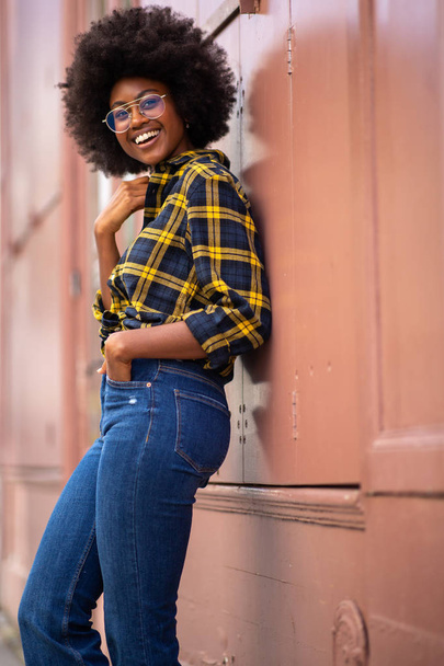 Zijportret van lachende jonge zwarte vrouw met afro haar leunend tegen de muur  - Foto, afbeelding