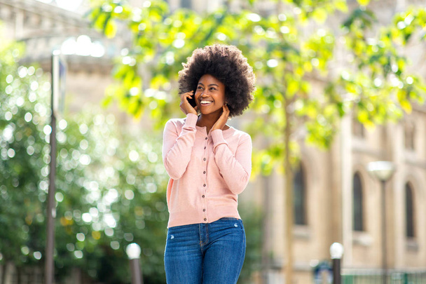 Portrét šťastně se usmívající krásné africké ženy kráčející a mluvící s mobilním telefonem venku ve městě - Fotografie, Obrázek