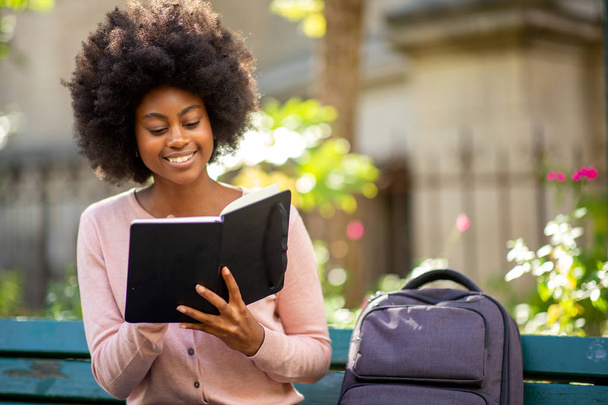 Portret młodej, uśmiechniętej Afroamerykanki czytającej książkę na zewnątrz - Zdjęcie, obraz