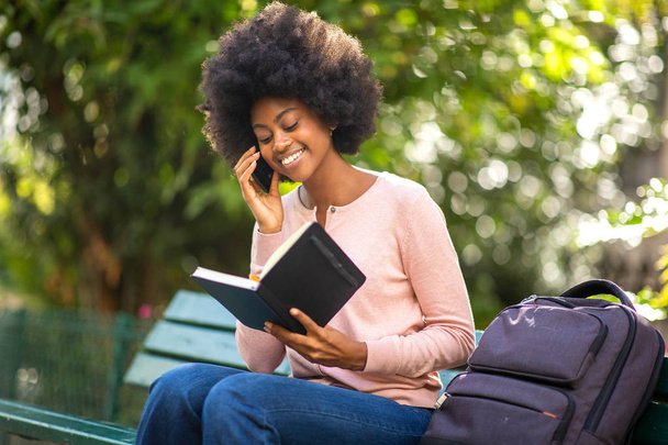 Portret van een vrolijke glimlachende zwarte vrouw buiten zittend met boekentas en pratend met mobiele telefoon - Foto, afbeelding