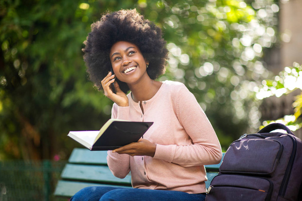 Πορτρέτο της χαμογελαστής μαύρης γυναίκας που κάθεται έξω με την τσάντα του βιβλίου και μιλάει με κινητό - Φωτογραφία, εικόνα