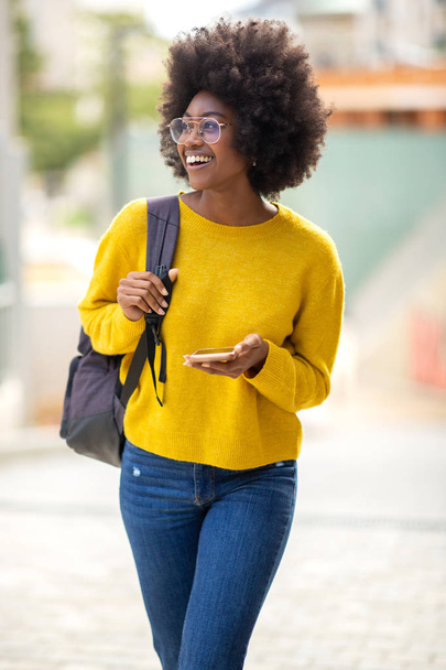 Portrait of smiling african american woman with glasses walking in city and looking at mobile phone - Foto, imagen