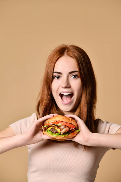Young red-haired girl holds a burger sandwich, is surprised, on a beige background - 写真・画像