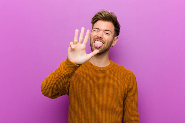 young man smiling and looking friendly, showing number four or fourth with hand forward, counting down against purple background - Foto, imagen