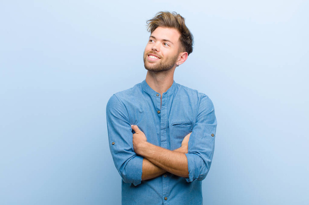 young businessman feeling happy, proud and hopeful, wondering or thinking, looking up to copy space with crossed arms against blue background - Photo, image