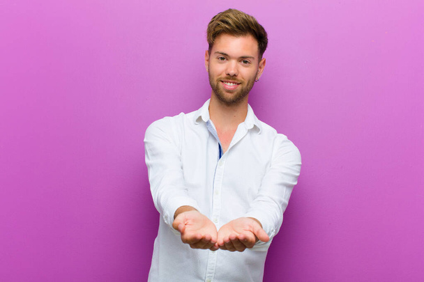 young man smiling happily with friendly, confident, positive look, offering and showing an object or concept against purple background - Fotoğraf, Görsel
