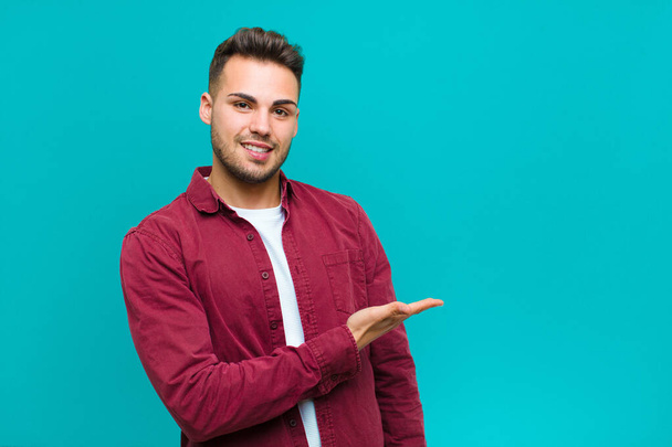 young hispanic man smiling cheerfully, feeling happy and showing a concept in copy space with palm of hand against blue background - Foto, Imagen