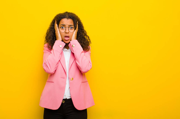 black business woman looking unpleasantly shocked, scared or worried, mouth wide open and covering both ears with hands against orange wall - Foto, imagen