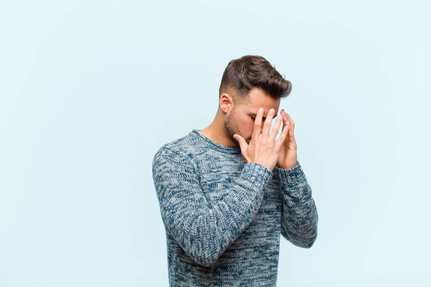 young hispanic man covering eyes with hands with a sad, frustrated look of despair, crying, side view against blue background - Foto, Imagem