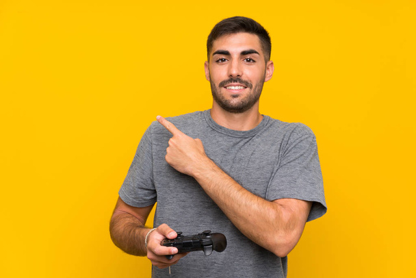 Young handsome man playing with a video game controller over isolated yellow background pointing to the side to present a product - Fotografie, Obrázek