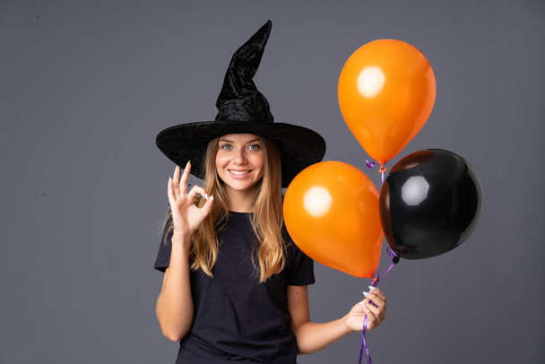 Young witch holding black and orange air balloons showing an ok sign with fingers - Foto, Imagen