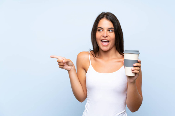 Elinde kahve tutan genç bir kadın izole edilmiş mavi arka planda şaşırmış ve parmağını kenarı işaret ediyor. - Fotoğraf, Görsel