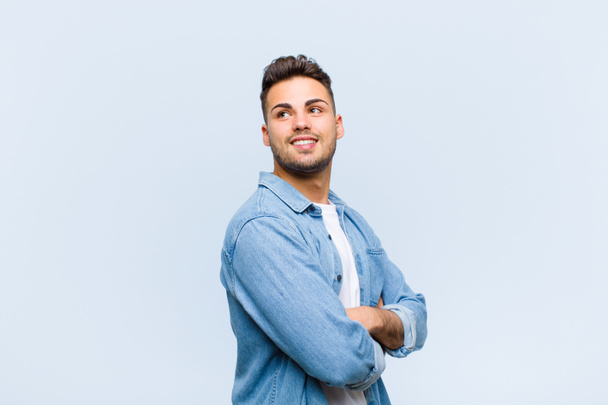 fiatal spanyol férfi vidáman mosolyog, boldognak, elégedettnek és nyugodtnak érzi magát, keresztbe tett karokkal és a kék fal felé néz - Fotó, kép