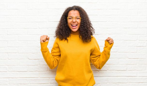 young black woman feeling happy, surprised and proud, shouting and celebrating success with a big smile against brick wall - 写真・画像