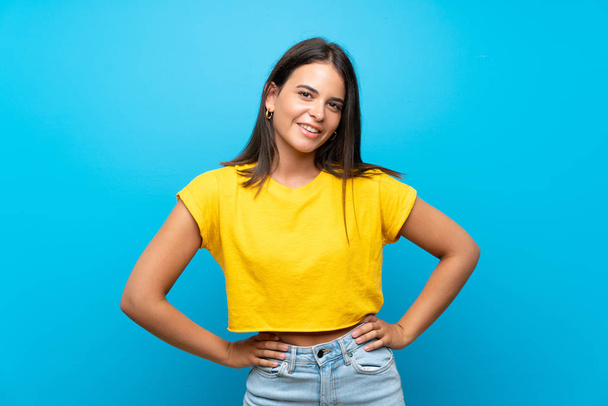Young girl over isolated blue background posing with arms at hip and smiling - Фото, зображення