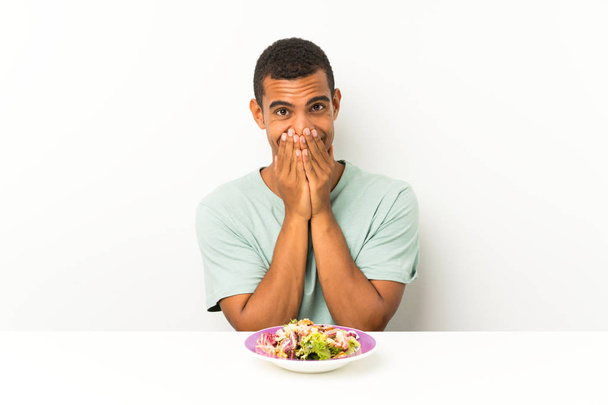 junger schöner Mann mit Salat an einem Tisch mit überraschendem Gesichtsausdruck - Foto, Bild