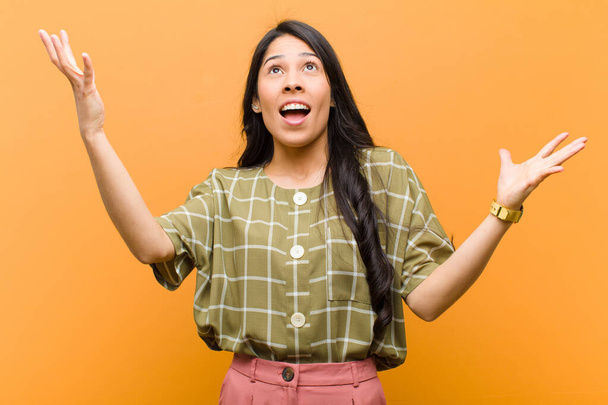 young pretty hispanic woman feeling happy, amazed, lucky and surprised, celebrating victory with both hands up in the air against brown wall - Foto, imagen