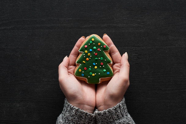 cropped view of woman holding Christmas tree cookie in hands on black wooden table - Foto, Imagem