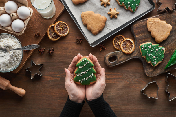 vista cortada da mulher segurando biscoito árvore de Natal perto de ingredientes e especiarias na mesa de madeira
 - Foto, Imagem