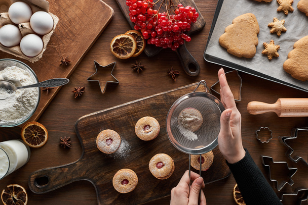 καλλιεργημένη άποψη της γυναίκας κοσκινίζοντας ζάχαρη σε σκόνη για τα χριστουγεννιάτικα μπισκότα στο ξύλινο τραπέζι - Φωτογραφία, εικόνα
