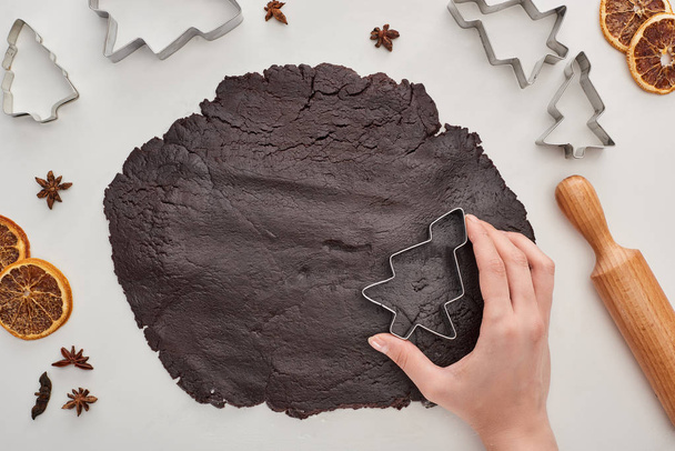 cropped view of woman holding Christmas tree dough mold on raw dough on white background near anise, dough molds, rolling pin and dried citrus - Valokuva, kuva